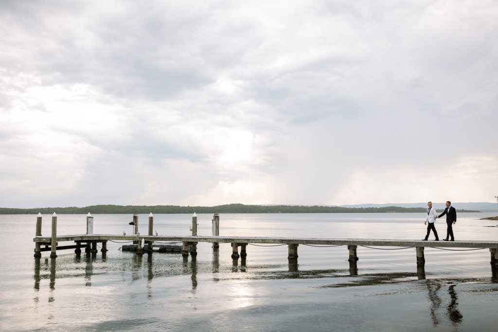 husband and husband holding hands walking on a jetty at raffertys resort during the groom photo shoot