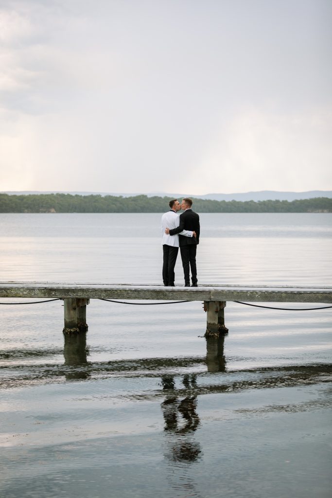 husband and husband holding hands walking on a jetty at raffertys resort during the groom photo shoot