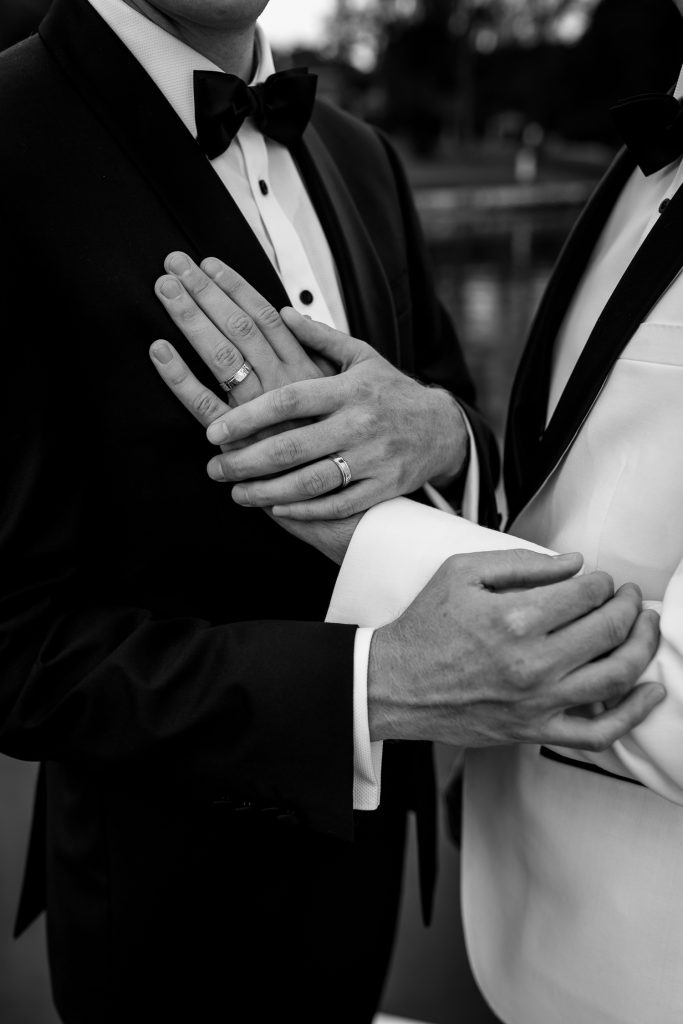husband and husband holding hands showing us their wedding rings at raffertys resort during the groom photo shoot