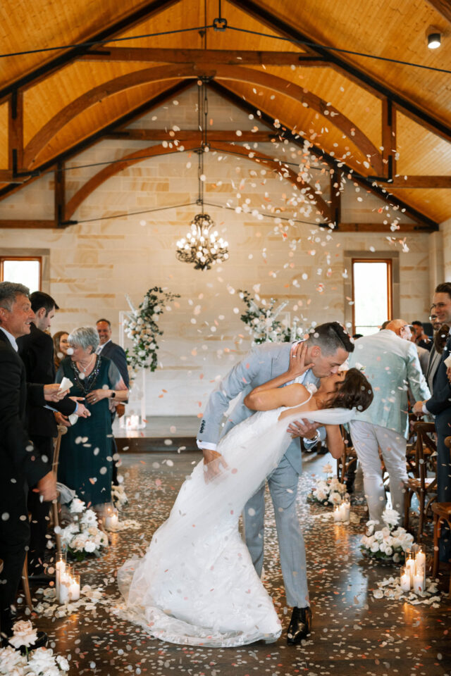 husband and wife dip and kiss in the isle of a chapel with confetti in the hunter valley